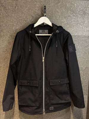 Marinblå vindjacka från Brixtol - Svart vindjacka från Brixtol med huva och två stora fickor framtill med knappstängning och även 2 st mindre innanför dem. Jackan har en snygg silverfärgad dragkedja och snörning vid huvan. Patch med Brixtol-logga på ärmen ger en cool touch som även är avtagbar. Perfekt för blåsiga dagar. Köpt för 1999kr för några månader sen bara så den är i topp skick!