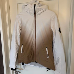 Vit och beige skidjacka från J.Lindeberg - Säljer en helt oanvänd skitsnygg J.Lindeberg Halo Pro skidjacka. Den är i nyskick och alla originaletiketter/lappar sitter kvar. - Vattentät (30k mm) och 4-vägs stretch för maximal komfort.  - Snölås, ventilationsblixtlås och liftkortsficka. Nypris : 7000kr. Hör av er vid frågor och för fler bilder 🫶