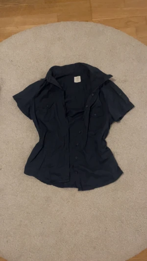 Mörkblå kortärmad skjorta med fickor - Svart skjorta i bomull med korta ärmar och klassisk krage. Skjortan har två bröstfickor med lock och knappar samt knäppning framtill. Perfekt för en avslappnad och stilren look. Har du några frågor eller vill ha mått är det bara att skriva till mig!💙💙