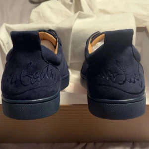 Mörkblå sneakers från Christian Louboutin - Snygga mörkblå sneakers i mocka från Christian Louboutin med den ikoniska röda sulan. Skorna har diskret snörning och broderad logotyp på hälen. Perfekta för att ge en lyxig touch till din stil.