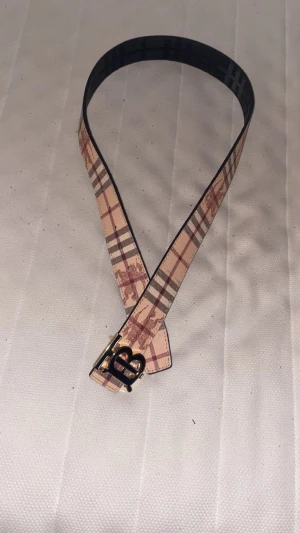 Burberry vändbart skärp med TB-spänne - Säljer ett vändbart Burberry-bälte med klassiskt rutigt mönster i beige, brun och röd på ena sidan och svart med diskret relief på andra. Snyggt TB-spänne i guld. Bältet är gjort i skinn och har en stilren, modern vibe som lyfter din outfit.