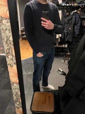 Mörkblå stickad tröja J.Lindeberg M - Snygg mörkblå stickad tröja från J.Lindeberg i storlek M. Tröjan har ett klassiskt kabelstickat mönster, rund hals och ribbade muddar vid ärmslut och nederkant. Perfekt för dig som gillar stilrena och tidlösa plagg.