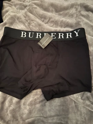 Svarta Burberry Storlek S-M - Snygga svarta shorts från Burberry med bred resår och tydlig vit logotyp på midjan. De har en klassisk, tight passform och är gjorda i mjukt syntetmaterial som känns skönt mot huden. Perfekta för dig som gillar stilrena och exklusiva märken. Storlek S-M