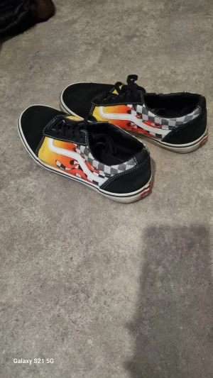 Vans Old Skool flames & checkerboard - Säljer ett par Vans Old Skool sneakers med svart tå och häl, klassisk vit sula och snörning. Sidorna har ett schackrutigt mönster i grått och vitt, samt coola orange och röda flames. Materialet är canvas och skinn, med rund tå och platt sula.