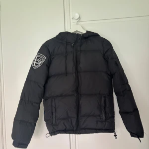 D.brand, svart dunjacka. - Dunjacka med rak passform från d.brand, fastsydd luva,dragkedjefickor framtill och en innerficka, d.brand sköld på höger ärm. Fyllning 90% dun och 10% fjäder. Säljer den eftersom jag har en ny vinterjacka. 