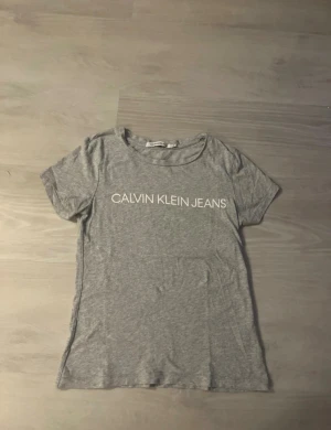 Grå t-shirt Calvin Klein Jeans XS - Snygg grå t-shirt från Calvin Klein Jeans i storlek XS. T-shirten har klassisk rund hals och korta ärmar, med vit logotyp framtill. Mjuk och tunn bomullskvalitet som känns skön mot huden. Perfekt för en chill och stilren look.