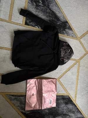 Svart hoodie från Syna med tryck - Svart hoodie från Syna med coolt graffiti-tryck på huvan. Tröjan har snörning vid huvan, stor magficka och ribbade muddar vid ärmslut och nederkant. Materialet är 80% bomull och 20% polyester, vilket gör den både mjuk och skön. Perfekt för dig som gillar streetwear.