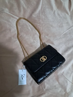 Svart quiltad axelväska RF Fashion Bag - Stilren svart axelväska med quiltad design och guldfärgad kedja. Väskan har ett snyggt guldspänne framtill och flera fack inuti, inklusive ett med dragkedja. Tillverkad i syntetmaterial med lyxig känsla och passar perfekt till din outfit.