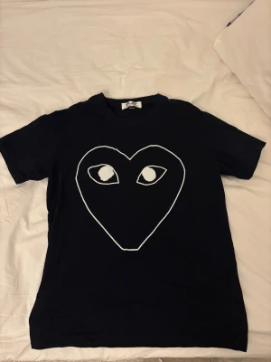  CDG tshirt - Marin blå t-shirt från Comme des Garçons Play med stort vitt hjärta och ögon tryckt på framsidan. Klassisk rund hals och korta ärmar. Materialet är mjuk bomull som känns skön mot huden. Perfekt för dig som gillar statement-plagg och ikoniska designer. Den är i storlek M men sitter mer som S. Tshirten är använd 2 gånger.