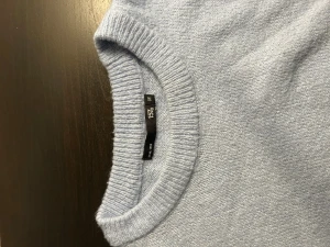 Ljusblå stickad tröja från 157 - Mysig ljusblå stickad tröja från 157 med rund hals och ribbade muddar. Perfekt för att hålla värmen under kyliga dagar. Enkel och stilren design som är lätt att matcha med dina favoritjeans eller kjol.