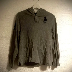 Grå hoodie från Polo Ralph Lauren - Grå hoodie från Polo Ralph Lauren med huva och långa ärmar. Tröjan har en stor svart polospelare broderad på bröstet och en svart siffra 3 på ena ärmen. Tillverkad i mjuk bomull, perfekt för chill dagar. Klassisk knappslå vid halsen och relaxed passform. 14-16