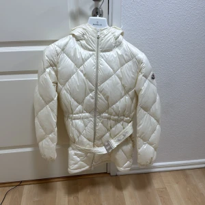 Vit quiltad dunjacka från Moncler - Säljer en snygg vit dunjacka från Moncler med huva och bälte i midjan. Jackan har ett quiltat mönster, dragkedja framtill och Moncler-logga på ärmen. Storlek M men passar S