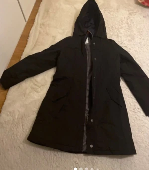 Svart parkajacka från Marccain, strl 38 - Svart parkajacka från Marko polo med huva och Thermore-isolering. Jackan har långa ärmar, knappar och dragkedja framtill samt stora fickor. Perfekt för kyliga dagar och har en stilren, enkel design.