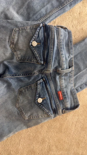Blå bootcut jeans från Divided - Säljer ett par blå bootcut jeans från Divided med klassiska fem fickor och slitna, fransiga benslut. Jeansen har en normal passform och är tillverkade i bomull. Perfekt för dig som gillar en avslappnad och retro vibe.