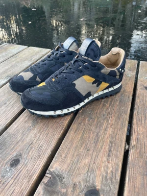Valentino Garavani Rockrunner sneakers - Snygga Valentino Garavani Rockrunner sneakers med mörkblå, beige och gul camouflage-mönster. Perfekta nu inför vintern. Ens skon saknar 3 nitar men det är inget som märks ser också ut att vara nånting på sulan på ena skon. Hör av dig vid frågor