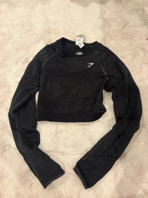 Svart croerppad långärmad topp Gymshark - Svart croppad långärmad topp från Gymshark med cool snörning i ryggen och diskret mönstrad struktur. Toppen har rund halsringning och är tillverkad i en stretchig blandning av nylon, polyester och elastan. 