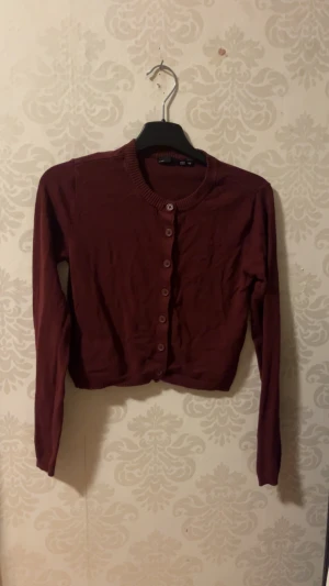 Vinröd kofta från 157, XS - En vinröd kofta med rund hals och knappar hela vägen framtill från 157. Modellen är croppad och har långa ärmar med ribbade muddar. Perfekt att slänga över en topp för en chill look.