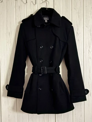 Svart trenchcoat med bälte från H&M - Stilren svart trenchcoat från H&M med dubbelknäppning, axelklaffar och bälte i midjan. Jackan har långa ärmar, klassisk krage och knappar vid ärmsluten. Den ger en snygg siluett och passar perfekt till kyligare dagar.