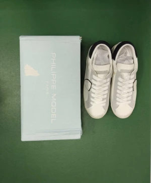 Philippe Model vita sneakers med svart detalj - Snygga vita sneakers från Philippe Model med svart mockadetalj på hälen och grå inslag på sidorna. Skorna har snörning, platt sula och rund tå. Tillverkade i skinn och mocka för en lyxig känsla. Box medföljer. Perfekt för dig som gillar stilrena och trendiga sneakers.