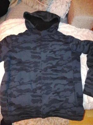 Camouflage teddyjacka från Denim Day - Mörkblå teddyjacka med camouflagemönster från Denim Day. Jackan har huva, dragkedja framtill och är fodrad med mjuk svart fleece. Perfekt för dig som gillar streetstyle och vill ha en varm jacka med cool look.