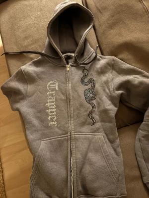 Grå rhinestone hoodie med orm från Trapper - Grå hoodie från Trapper i storlek S med dragkedja och huva. På framsidan finns ett coolt ormtryck och text i glittriga stenar. Tröjan har snörning vid huvan och är tillverkad i mjukt fleecematerial. Perfekt för dig som gillar streetstyle och detaljer. Använd väldigt få gånger