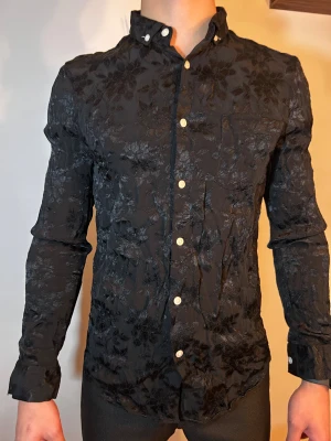 Svart blommig skjorta med knappar - Svart skjorta med blommigt mönster i glansigt tyg och vita knappar framtill. Skjortan har lång ärm, klassisk krage med knappdetaljer och en slim passform som sitter snyggt på kroppen. Perfekt för dig som vill sticka ut lite extra.