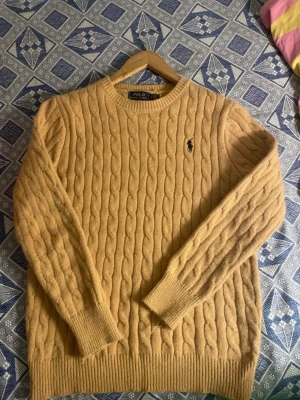 Polo Ralph Lauren Cable Knit Sweater - Denna klassiska kabelstickade tröja från Polo Ralph Lauren är ett måste i varje garderob. Tröjan är tillverkad av mjukt, bekvämt material och har en tidlös design som passar perfekt för både vardag och fest. 