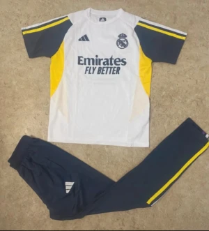 Real Madrid träningsset Adidas - Säljer ett Real Madrid träningsset från Adidas med vit t-shirt och mörkblå byxor. Tröjan har gula och mörkblå detaljer samt klubbmärke och sponsortryck framtill. Byxorna är mörkblå med gula ränder längs sidorna och Adidas-logga på benet. Perfekt för fotbollsträning.