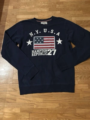 Mörkblå sweatshirt Hampton Republic - Mörkblå sweatshirt från Hampton Republic med stort tryck av amerikansk flagga och texten N.Y.U.S.A samt Hampton Republic 27 på bröstet. Tröjan har långa ärmar, ribbade muddar och rund hals med rödvit randig insida. Perfekt för dig som gillar sportig och casual stil.