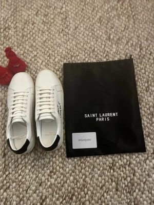 Saint Laurent vita sneakers i skinn - Snygga vita Saint Laurent sneakers med svart detalj på hälen och handskriven logga på sidan. Skorna är i slätt skinn med platt, beige sula och klassisk snörning. Perfekta för dig som gillar stilrena och exklusiva sneakers med en lyxig touch. St-43 passar även 42. Skick 10/10 priset går att ändras. 