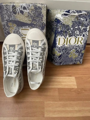  Dior sneakers  - Säljer ett par snygga Dior sneakers i grått och vitt med klassiskt Dior-monogram på sidan. Skorna har snörning, platt sula och rund tå. Märket är tydligt på både plösen och sulan. Materialet är textil och syntet, vilket ger en lyxig känsla och cool look. ÄKTA!! Fix de i present o h därför har inte kvittot men har boxen och allt annat man får med! Pris kan diskuteras vid snabba köp! Helt oanvända 