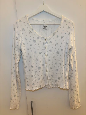 Vit blommig kofta från Censored - Supersöt vit kofta från Censored med små blå blommor och ribbad struktur. Koftan har långa ärmar, knappar framtill och vågig kant nertill. Perfekt för vår eller sommar och ger en romantisk vibe till din outfit.