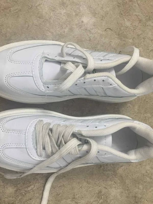 Vita Adidas sneakers strl 39 - Fräscha vita sneakers från Adidas med klassiska ränder på sidan och rund tå. Skorna har snörning och en platt sula i vitt. Ovandelen är i syntetmaterial med detaljerade sömmar och vadderad krage för extra komfort. Perfekt för dig som gillar stilrena och sportiga skor.