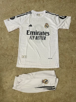 Real Madrid Mbappé matchställ - Säljer ett Real Madrid matchställ med Mbappé och nummer 9 på ryggen. Setet består av vit tröja och shorts med svarta detaljer och klubbens emblem. Adidas logga och 'Emirates Fly Better' tryck på framsidan. Perfekt för fotbollsfans som vill ha en snygg och autentisk look.