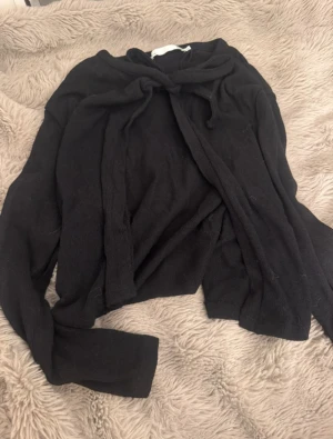 Svart stickad kofta med knyt - Svart stickad kofta med långa ärmar och snyggt knyt framtill. Perfekt att slänga över en t-shirt eller linne för en chill och stilren look. Mjuk och skön känsla, passar dig som gillar enkla och klassiska plagg.