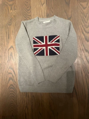 Grå stickad tröja med Union Jack - Grå stickad tröja från Sfera med lång ärm och ribbade muddar. På bröstet finns en stor Union Jack-flagga i marinblått, vinrött och vitt. Rund hals och klassisk passform, perfekt för dig som gillar coola detaljer och brittisk stil.