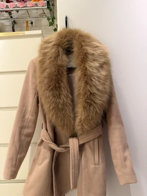 Beige kappa med päls från Chiara Forthi - Snygg beige kappa från Chiara Forthi med stor fluffig pälsimitation runt kragen och bälte i midjan. Jackan har en klassisk, elegant look och är helfodrad med ljus insida. Perfekt för dig som vill ha en chic vibe i höst och vinter.