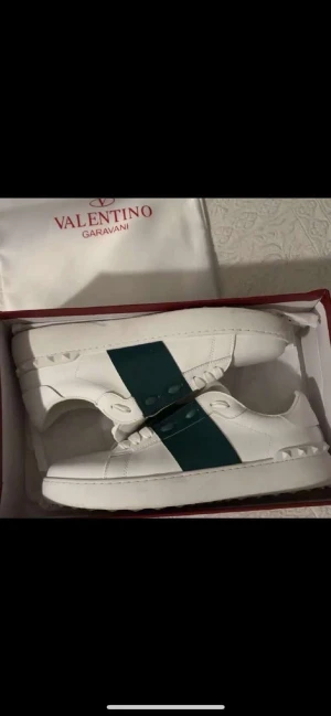 Valentino Garavani Open sneakers vit/grön - Säljer ett par Valentino Garavani Open sneakers i vitt kalvskinn med bred grön rem över mitten. Skorna har klassisk låg profil, snörning och coola nitar på hälen. Snygg och stilren design som sticker ut med den gröna detaljen. Box och dustbag medföljer. Priset  kan diskuteras 🥂
