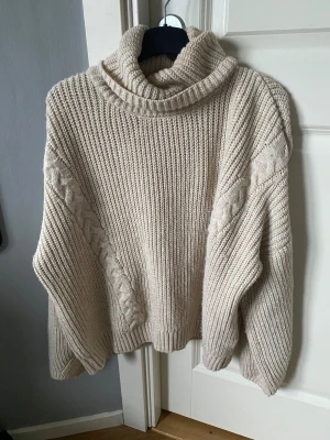 Beige stickad polotröja med flätmönster - Säljer en beige stickad polotröja med bred krage och snygga flätade detaljer längs ärmarna. Tröjan har en loose passform och är ribbstickad för extra mysig känsla. Perfekt för kyliga dagar när du vill vara både bekväm och stilren.