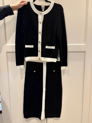 Svart och vit stickad dräkt - Snygg tvådelad dräkt med svart kjol i M och matchande kofta i S, båda med vita kontrastdetaljer. Koftan har rund hals, silverfärgade knappar och två fickor framtill. Kjolen är rak med vita kanter och dekorativa fickor med knappar och ett litet sprund framtill. Materialet är stickat och ger en elegant look.