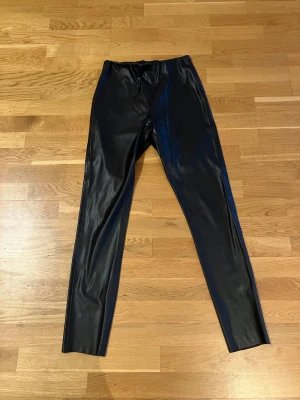 Svarta leggings i skinnimitation - Säljer ett par svarta leggings i glansig skinnimitation. De har en tight passform och elastisk midja som gör dem riktigt bekväma. Perfekta för dig som vill ha en edgy och trendig look. Passar till oversized tröjor eller crop tops.
