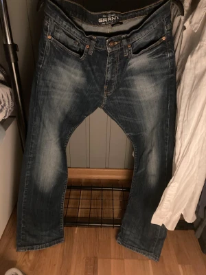 Grant 405 blå jeans W32 L30 - Säljer ett par snygga Grant 405 jeans i klassisk blå tvätt med slitningar och kontrastsömmar. Modellen har raka ben, fem fickor och läderpatch bak i midjan. Perfekt passform för dig som gillar en normal look och vill ha jeans som funkar till det mesta.