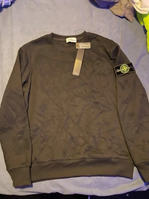 Svart Stone Island sweatshirt - Svart sweatshirt från Stone Island med klassisk rund hals och ribbade muddar. Ikonisk avtagbar patch med logga på vänster ärm. Tröjan är i mjuk bomull och har en clean, stilren look som passar perfekt till streetwear.