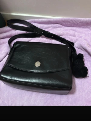 Svart axelväska i faux skinn med nalle - 🎀frågor detaljbild prisjustering pm Snygg svart axelväska  i faux skinn med justerbar rem och magnetlås. Väskan har en stilren, rektangulär form och ett fack med dragkedja på insidan. På sidan hänger en gullig svart nalle som detalj. Perfekt för dig som gillar klassisk stil med en twist.