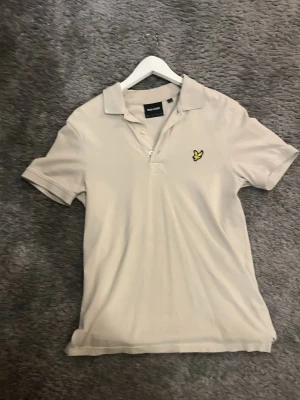Beige pikétröja från Lyle & Scott - Säljer en stilren beige pikétröja från Lyle & Scott med klassisk krage, korta ärmar och knäppning framtill. Tröjan har den ikoniska gula örnloggan broderad på bröstet och är tillverkad i bomull för en skön känsla. Passar dig mellan 170-180