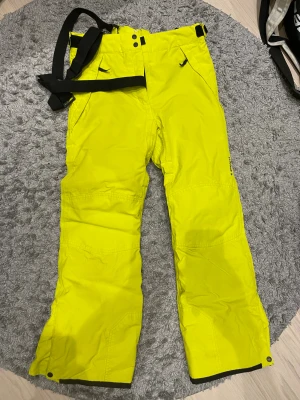 Gula Everest täckbyxor med hängslen - Säljer ett par sjukt snygga täckbyxor från Stadium i gul  färg. De har svart bälte, praktiska fickor med dragkedja och förstärkta knän. Perfekta för vintern och skidåkning, med bred passform och slitstarkt material. Inköpta förra vintern från stadium och använda ett fåtal gånger. Bra kvalitet dessutom. 