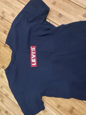 Svart Levi's t-shirt med röd logga - Snygg svart t-shirt från Levi's med klassisk röd logga tryckt på bröstet. T-shirten har rund hals och korta ärmar, perfekt för en avslappnad och cool stil. Tillverkad i mjuk bomull som känns skön mot huden.