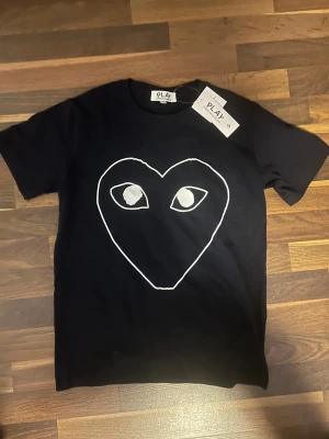 Svart Comme des Garçons Play t-shirt - Svart t-shirt från Comme des Garçons Play med ett stort vitt hjärta med ögon tryckt på framsidan. Klassisk rund hals och korta ärmar. Tillverkad i mjuk bomull för en skön känsla. Perfekt statement-plagg för dig som gillar streetstyle.