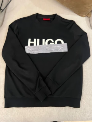Svart sweatshirt från HUGO - Säljer en svart sweatshirt från HUGO med stor vit logga framtill och en grå detalj över bröstet. Tröjan har rund halsringning och långa ärmar. Perfekt för dig som gillar stilrena och trendiga plagg med en twist.