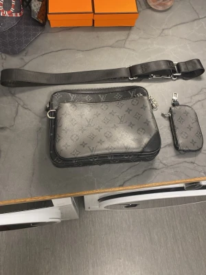 Louis Vuitton svart axelväska monogram - Snygg svart axelväska från Louis Vuitton med klassiskt monogrammönster i grått. Väskan har justerbar axelrem, flera fack med dragkedja och en matchande liten pouch som kan fästas. Tillverkad i skinn med silverfärgade detaljer.